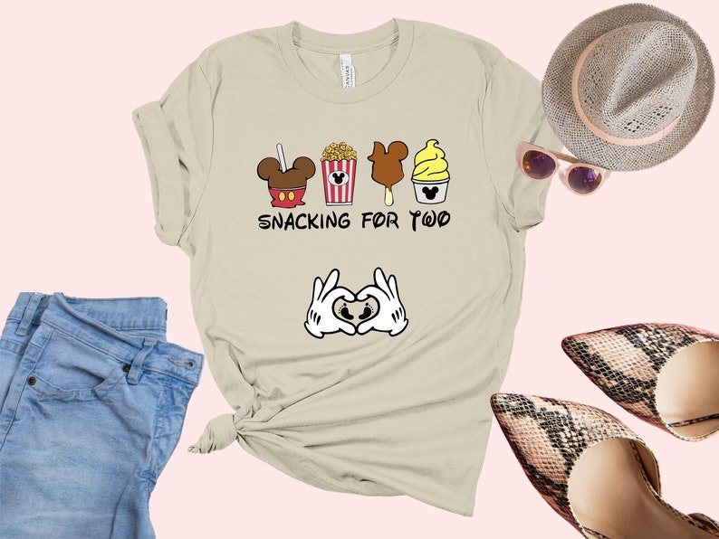 May include: A light beige t-shirt with a graphic of Mickey Mouse ears, popcorn, a Mickey Mouse ice cream bar, and a Mickey Mouse ice cream cone. The text "Snacking for Two" is printed below the graphic. Two white Mickey Mouse hands are making a heart shape with their fingers below the text.