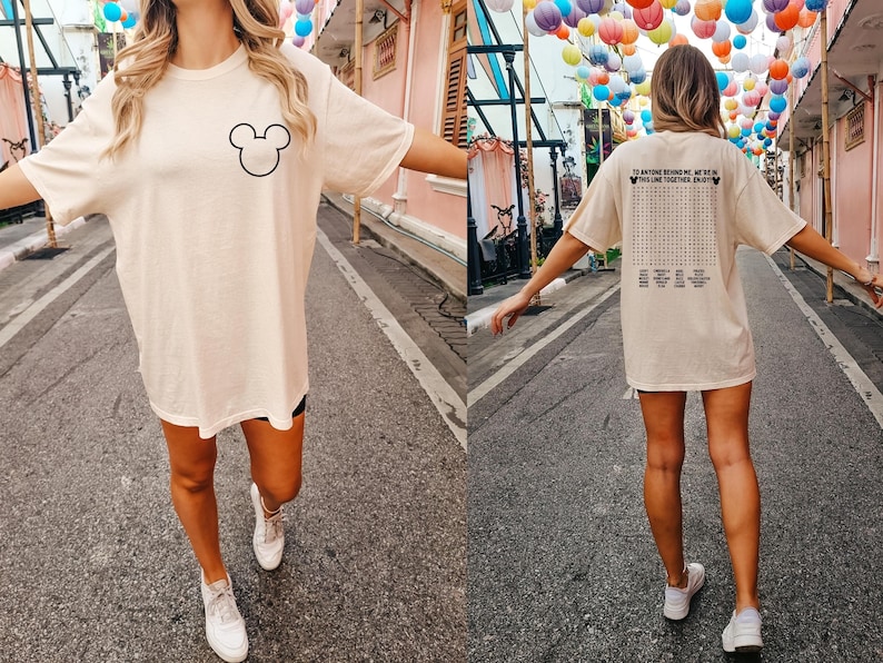 Pode incluir: Camiseta creme oversized com um contorno preto da cabe&ccedil;a do Mickey Mouse na frente. A parte de tr&aacute;s da camisa tem um jogo de palavras com o texto "TO ANYONE BEHIND ME, WE'RE IN THE LINE TOGETHER, ENJOY".