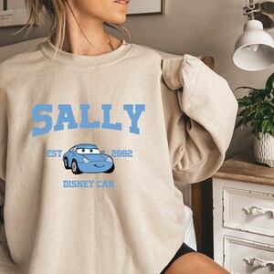 May include: A light beige sweatshirt with a blue graphic print of a cartoon car with the text "SALLY" above the car, "EST 2002" above the car, and "DISNEY CAR" below the car.