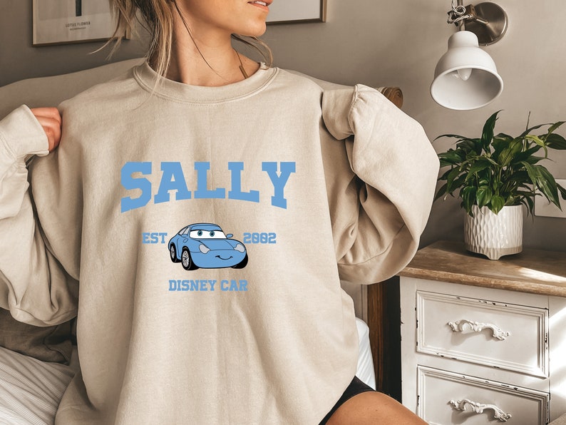 May include: A light beige sweatshirt with a blue graphic print of a cartoon car with the text "SALLY" above the car, "EST 2002" above the car, and "DISNEY CAR" below the car.