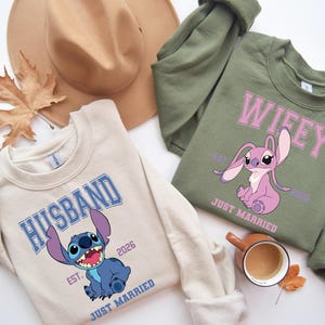 May include: Two sweatshirts, one beige and one olive green, with cartoon character graphics. The beige sweatshirt says "HUSBAND" and features a blue character. The green sweatshirt says "WIFEY" and features a pink character. Both say "EST. 2026" and "JUST MARRIED".