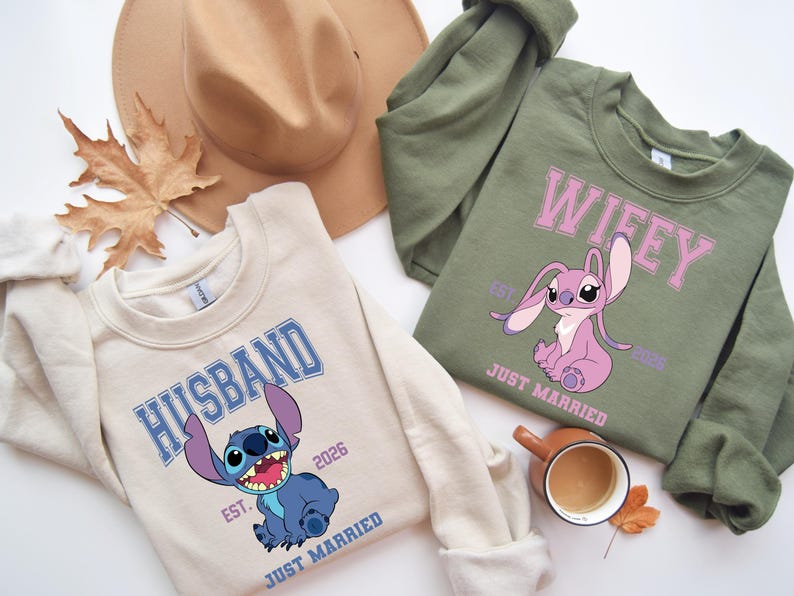 May include: Two sweatshirts, one beige and one olive green, with cartoon character graphics. The beige sweatshirt says "HUSBAND" and features a blue character. The green sweatshirt says "WIFEY" and features a pink character. Both say "EST. 2026" and "JUST MARRIED".