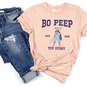 May include: A pink t-shirt with a blue and white illustration of Bo Peep from Toy Story. The text "Bo Peep" is in blue with the text "Est. 1999" and "Toy Story" in navy blue.