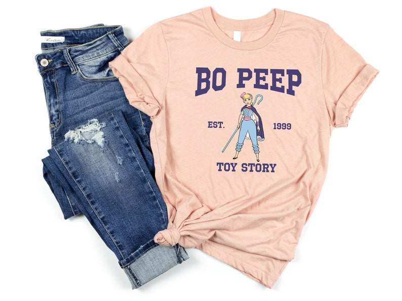 May include: A pink t-shirt with a blue and white illustration of Bo Peep from Toy Story. The text "Bo Peep" is in blue with the text "Est. 1999" and "Toy Story" in navy blue.