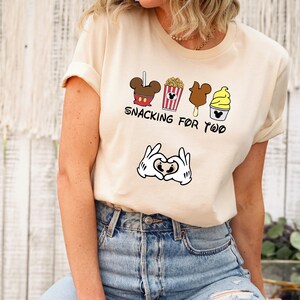 May include: A beige t-shirt with a graphic of Mickey Mouse snacks and the text "Snacking for Two". The graphic also includes a pair of Mickey Mouse hands making a heart shape with a baby footprint inside.