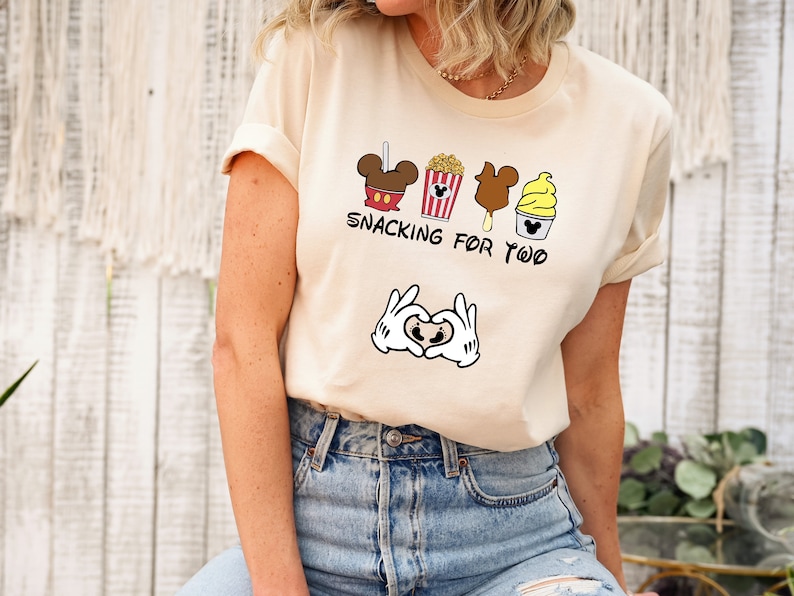May include: A beige t-shirt with a graphic of Mickey Mouse snacks and the text "Snacking for Two". The graphic also includes a pair of Mickey Mouse hands making a heart shape with a baby footprint inside.