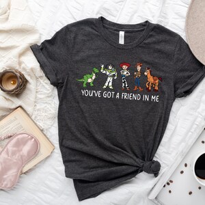 May include: A dark gray t-shirt with a white graphic of characters from the Toy Story movie franchise. The graphic includes Woody, Buzz Lightyear, Rex, and a horse. The text "You've got a friend in me" is printed below the graphic.