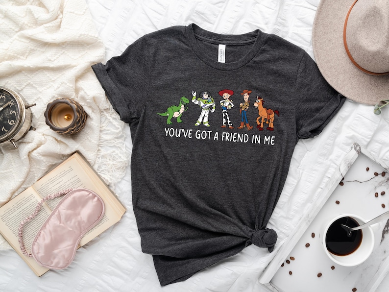 May include: A dark gray t-shirt with a white graphic of characters from the Toy Story movie franchise. The graphic includes Woody, Buzz Lightyear, Rex, and a horse. The text "You've got a friend in me" is printed below the graphic.