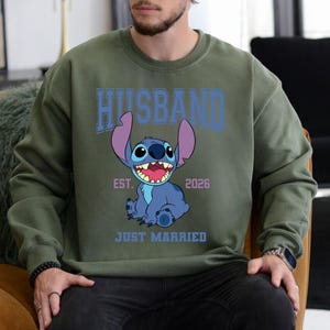 May include: Olive green sweatshirt with the word "HUSBAND" in blue block letters above a cartoon image of Stitch. The image includes the text "EST. 2026" and "JUST MARRIED" below the character.