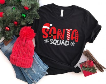 May include: Black t-shirt with a red and white Santa Claus graphic that says "Santa Squad". The shirt is laying on a pair of blue jeans and a red knit hat with a white pom-pom.