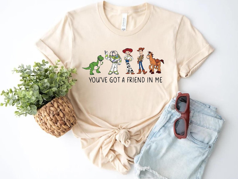 May include: A beige t-shirt with a graphic of the Toy Story characters Rex, Buzz Lightyear, Woody, Jessie, and Bullseye. The text "You've got a friend in me" is printed below the graphic.