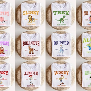 May include: Twelve white t-shirts with different Toy Story characters printed on them. Each shirt has the character's name, "Toy Story", and "EST." followed by a year.