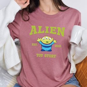 May include: A pink t-shirt with a green alien character from Toy Story. The shirt has the text "ALIEN EST. 2010 TOY STORY" printed on it.