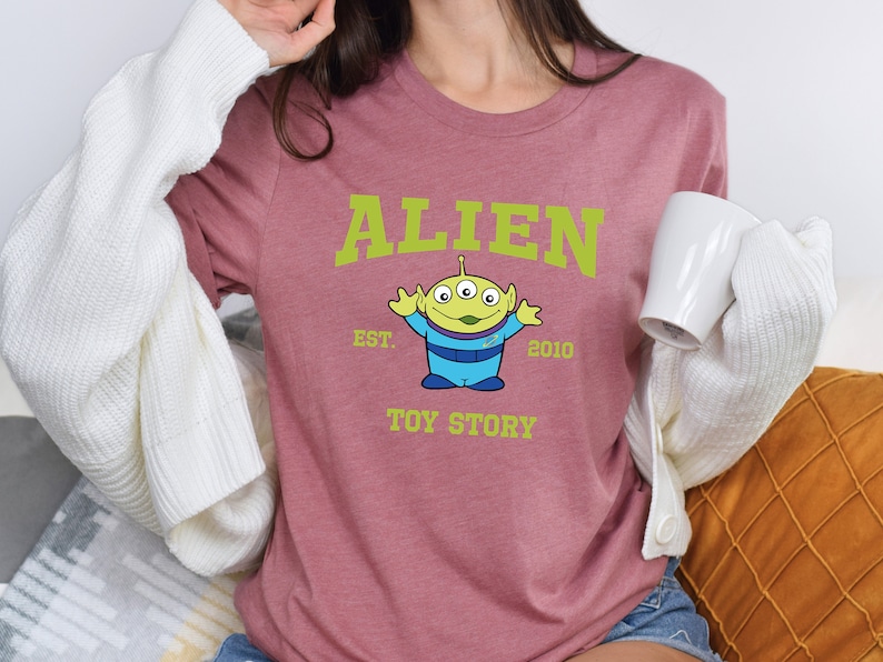 May include: A pink t-shirt with a green alien character from Toy Story. The shirt has the text "ALIEN EST. 2010 TOY STORY" printed on it.
