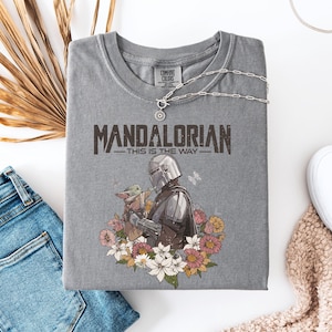 May include: Gray t-shirt featuring the text "MANDALORIAN - THIS IS THE WAY" with a graphic of the Mandalorian and Grogu surrounded by flowers. The shirt is paired with blue jeans, white sneakers, and a silver necklace.