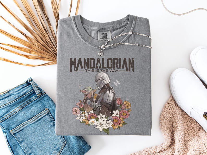 May include: Gray t-shirt featuring the text "MANDALORIAN - THIS IS THE WAY" with a graphic of the Mandalorian and Grogu surrounded by flowers. The shirt is paired with blue jeans, white sneakers, and a silver necklace.