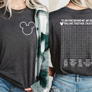 Pode incluir: Camiseta cinza escuro com um contorno branco do Mickey Mouse na frente. A parte de tr&aacute;s da camisa apresenta um quebra-cabe&ccedil;a de palavras com palavras tem&aacute;ticas da Disney e o texto "TO ANYONE BEHIND ME, WE'RE IN THIS LINE TOGETHER. ENJOY!"