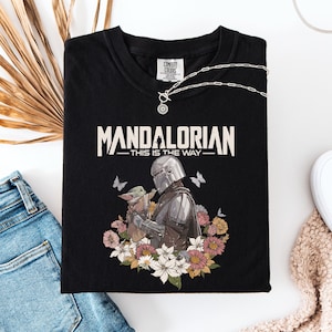 May include: Black t-shirt featuring the text "MANDALORIAN THIS IS THE WAY" in white. The graphic includes the Mandalorian and Grogu surrounded by flowers and butterflies. A silver necklace and white sneakers are also visible.