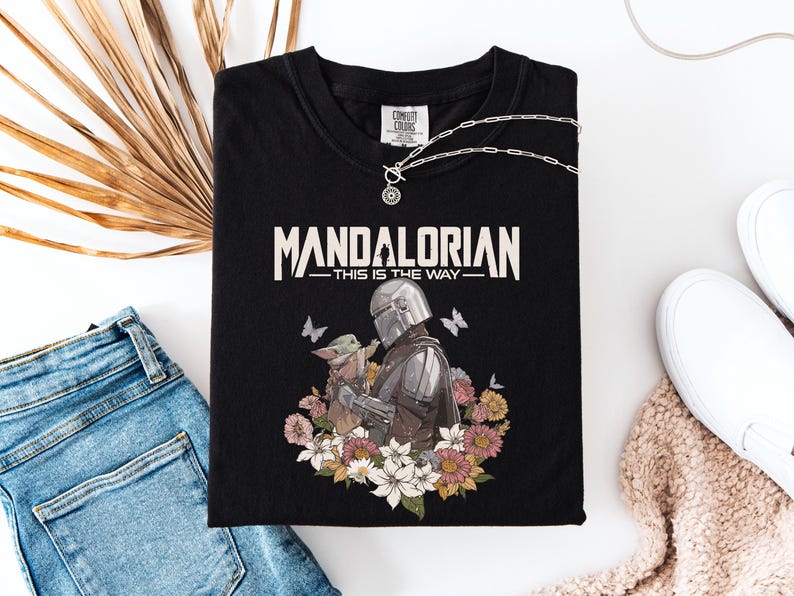 May include: Black t-shirt featuring the text "MANDALORIAN THIS IS THE WAY" in white. The graphic includes the Mandalorian and Grogu surrounded by flowers and butterflies. A silver necklace and white sneakers are also visible.