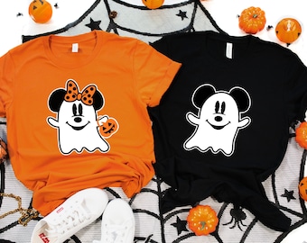 May include: Two Halloween-themed t-shirts. One is orange with a Minnie Mouse ghost design holding a pumpkin. The other is black with a Mickey Mouse ghost design. Both shirts have a white ghost outline.