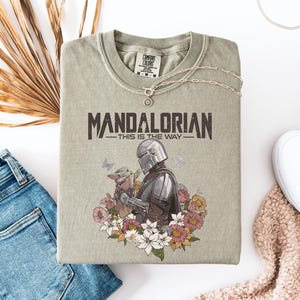May include: A sage green t-shirt featuring the text "MANDALORIAN - THIS IS THE WAY" in a distressed font. The graphic includes the Mandalorian and Grogu surrounded by flowers and butterflies. A silver necklace and white sneakers are also visible.
