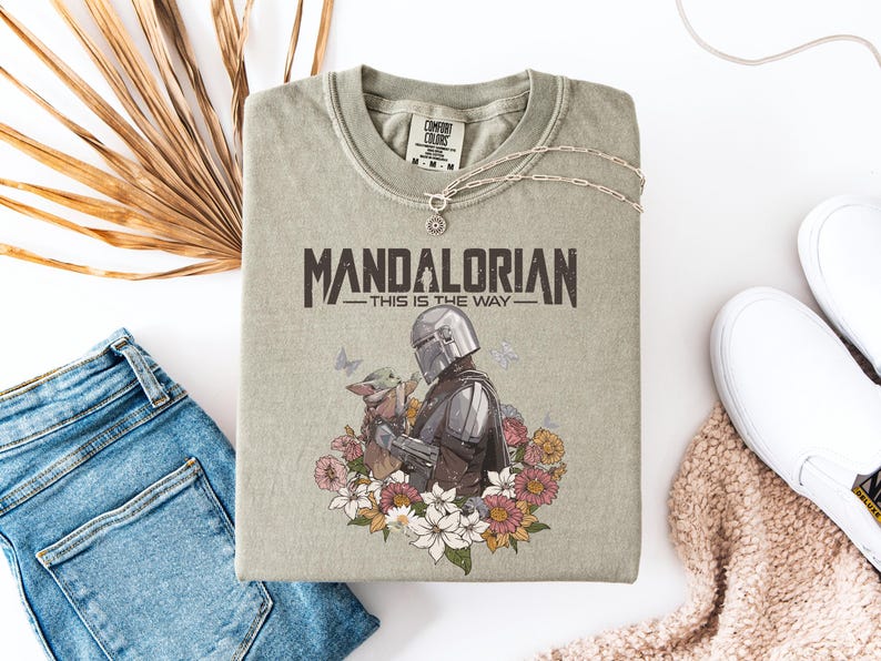 May include: A sage green t-shirt featuring the text "MANDALORIAN - THIS IS THE WAY" in a distressed font. The graphic includes the Mandalorian and Grogu surrounded by flowers and butterflies. A silver necklace and white sneakers are also visible.