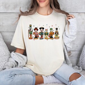 May include: Cream-colored t-shirt featuring a colorful graphic of Disney characters as nutcrackers. The design includes Mickey Mouse, Minnie Mouse, Donald Duck, and Goofy, each in festive outfits. The shirt is a comfortable, casual style.
