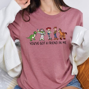 May include: A pink t-shirt with a graphic of characters from the Toy Story movie franchise. The graphic includes Woody, Buzz Lightyear, Rex, and Bullseye. The text "You've got a friend in me" is printed below the graphic.