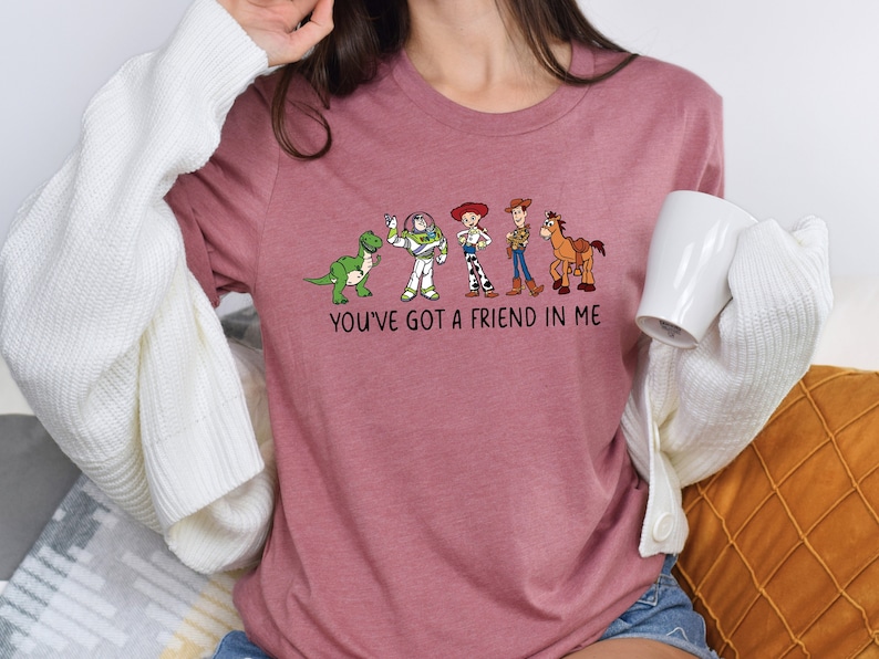 May include: A pink t-shirt with a graphic of characters from the Toy Story movie franchise. The graphic includes Woody, Buzz Lightyear, Rex, and Bullseye. The text "You've got a friend in me" is printed below the graphic.