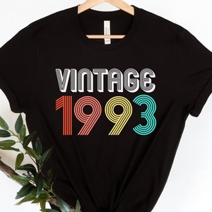May include: A black short-sleeved t-shirt featuring the word VINTAGE in white text above the year 1993. The numbers are displayed in a multi-line font with the number 1 in orange, 9 in pink, 9 in yellow, and 3 in teal, all on a black background.