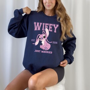 May include: Navy blue sweatshirt with the word "Wifey" in pink block letters above a pink cartoon character. Below the character are the words "Just Married" and the year "2026".