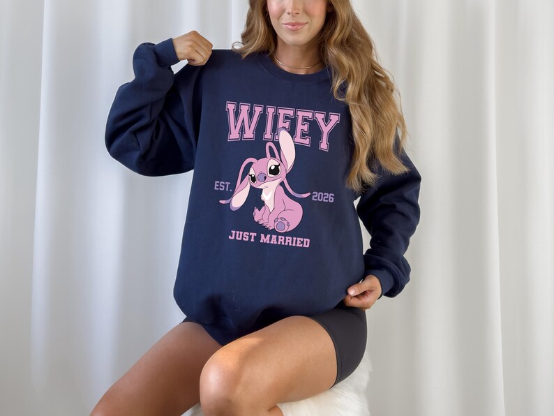 May include: Navy blue sweatshirt with the word "Wifey" in pink block letters above a pink cartoon character. Below the character are the words "Just Married" and the year "2026".