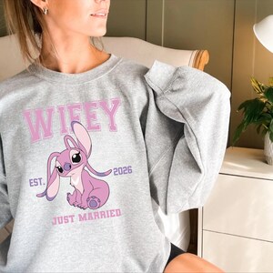May include: A light gray sweatshirt featuring a pink cartoon character and the word "Wifey" in pink block letters. Below the character are the words "Just Married" and the year "2026". The sweatshirt has long sleeves.