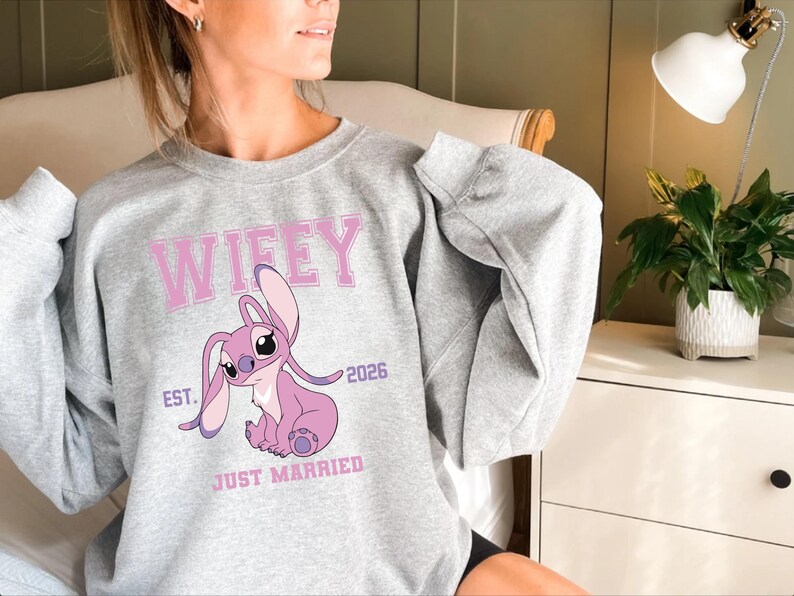 May include: A light gray sweatshirt featuring a pink cartoon character and the word "Wifey" in pink block letters. Below the character are the words "Just Married" and the year "2026". The sweatshirt has long sleeves.