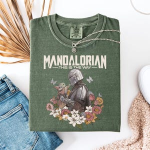 May include: Green t-shirt with the text "MANDALORIAN - THIS IS THE WAY" and a graphic of the Mandalorian and Grogu surrounded by flowers and butterflies. The shirt is laid flat with blue jeans, white shoes, and a necklace.