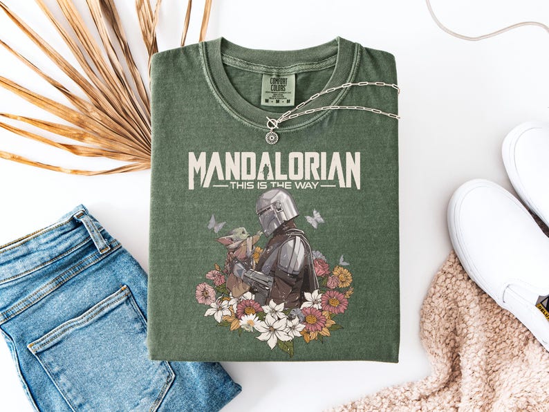 May include: Green t-shirt with the text "MANDALORIAN - THIS IS THE WAY" and a graphic of the Mandalorian and Grogu surrounded by flowers and butterflies. The shirt is laid flat with blue jeans, white shoes, and a necklace.