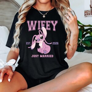 May include: Black t-shirt with the word "Wifey" in pink block letters above a pink cartoon character. Below the character are the words "Est. 2026" and "Just Married".