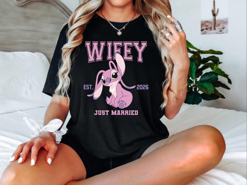 May include: Black t-shirt with the word "Wifey" in pink block letters above a pink cartoon character. Below the character are the words "Est. 2026" and "Just Married".