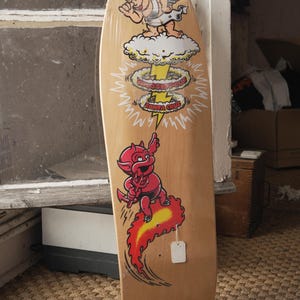 Puede incluir: Una patineta de madera con un dibujo animado de un ángel, una nube de hongo y un diablo. El ángel está en la parte superior, la nube de hongo en el medio y el diablo en la parte inferior. La patineta tiene una etiqueta de precio en blanco.