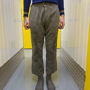 May include: Olive green striped pants with distressed details. The pants have a button closure and pockets. The person is wearing a navy blue long-sleeve shirt with colorful stripes on the wrist and gray socks.