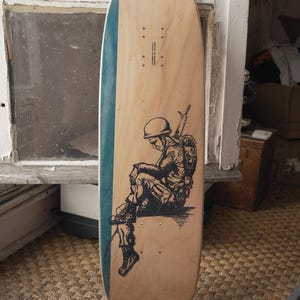 May include: A wooden skateboard with a teal stripe along the side. The board features a black and white illustration of a soldier sitting with their head in their hands. The skateboard has a natural wood finish.