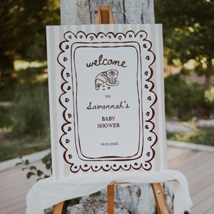 May include: A welcome sign for a baby shower, displayed on a wooden easel. The sign has a cream background with vertical stripes and a brown decorative border. The text reads "welcome to Savannah's Baby Shower" with the date 06.20.202X.