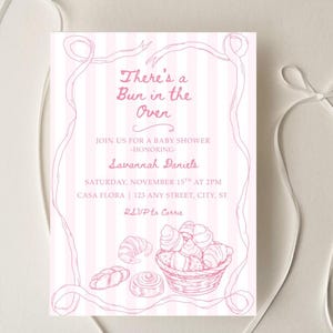 May include: Baby shower invitation with pink and white stripes, featuring the text "There's a Bun in the Oven". The invitation includes details for Savannah Daniels' baby shower, with a basket of baked goods illustration.