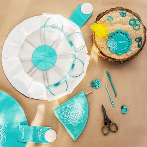 Peut inclure: Une boîte ronde à aiguilles à tricoter bleu sarcelle avec un intérieur blanc et une étiquette blanche qui dit "The Mindful Collection". La boîte est ouverte et montre les aiguilles à l'intérieur. Il y a aussi des marqueurs de points bleu sarcelle, un ruban à mesurer bleu sarcelle, une paire de ciseaux et une pochette bleu sarcelle avec un motif de mandala blanc.