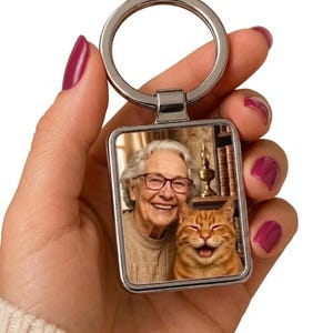 May include: A rectangular metal keychain featuring a printed photograph of an older person with white hair and glasses smiling next to an orange tabby cat with its eyes closed and mouth open in a happy expression, attached to a silver ring.