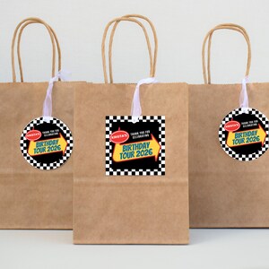 May include: Three brown paper gift bags with white ribbon handles. Each bag has a tag with a black and white checkered border and the text "Krista's Birthday Tour 2026". The tags are attached to the bags with white ribbon.