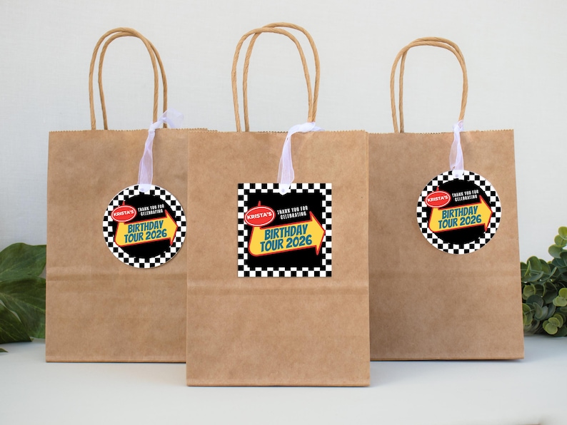 May include: Three brown paper gift bags with white ribbon handles. Each bag has a tag with a black and white checkered border and the text "Krista's Birthday Tour 2026". The tags are attached to the bags with white ribbon.