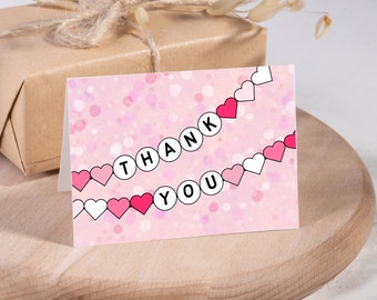 May include: A pink greeting card with the words "THANK YOU" in a heart-shaped design. The card has a pink background with white and pink confetti. A wrapped gift is in the background.