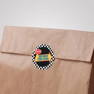 May include: A brown paper bag with a circular sticker. The sticker has a black background with a checkered border. The sticker text reads "Birthday Tour 2026" with an arrow design.