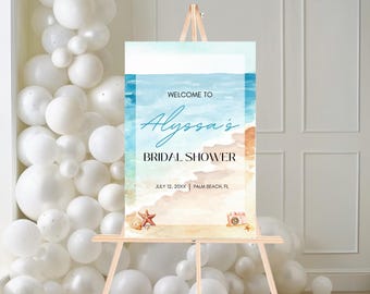 May include: A watercolor-style bridal shower welcome sign on a wooden easel. The sign features a beach scene with the text "Welcome to Alyssa's Bridal Shower" and the date and location. Decorative elements include a starfish and a camera.